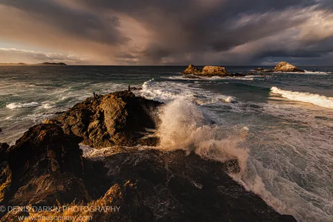 A wave explodes against a dark headland at Byron Bay, sending a tall plume of spray skyward while figures stand on the rocky outcrop.