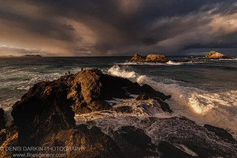 Rough, wet rock formations in low winter sunlight at Byron Bay, with storm clouds massing overhead and sea spray lifting from the surf below.
