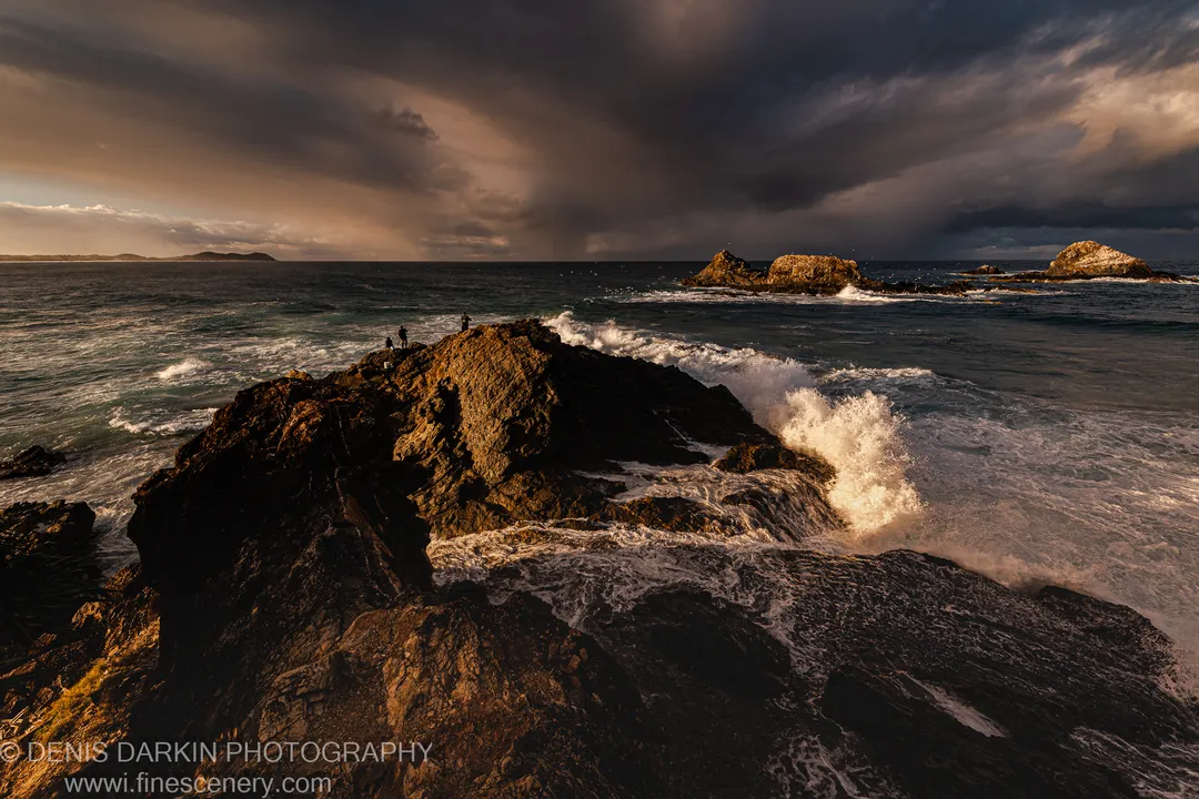 Small figures stand on a rugged coastal ridge at Byron Bay as sunlight breaks through storm cloud above a turbulent ocean.