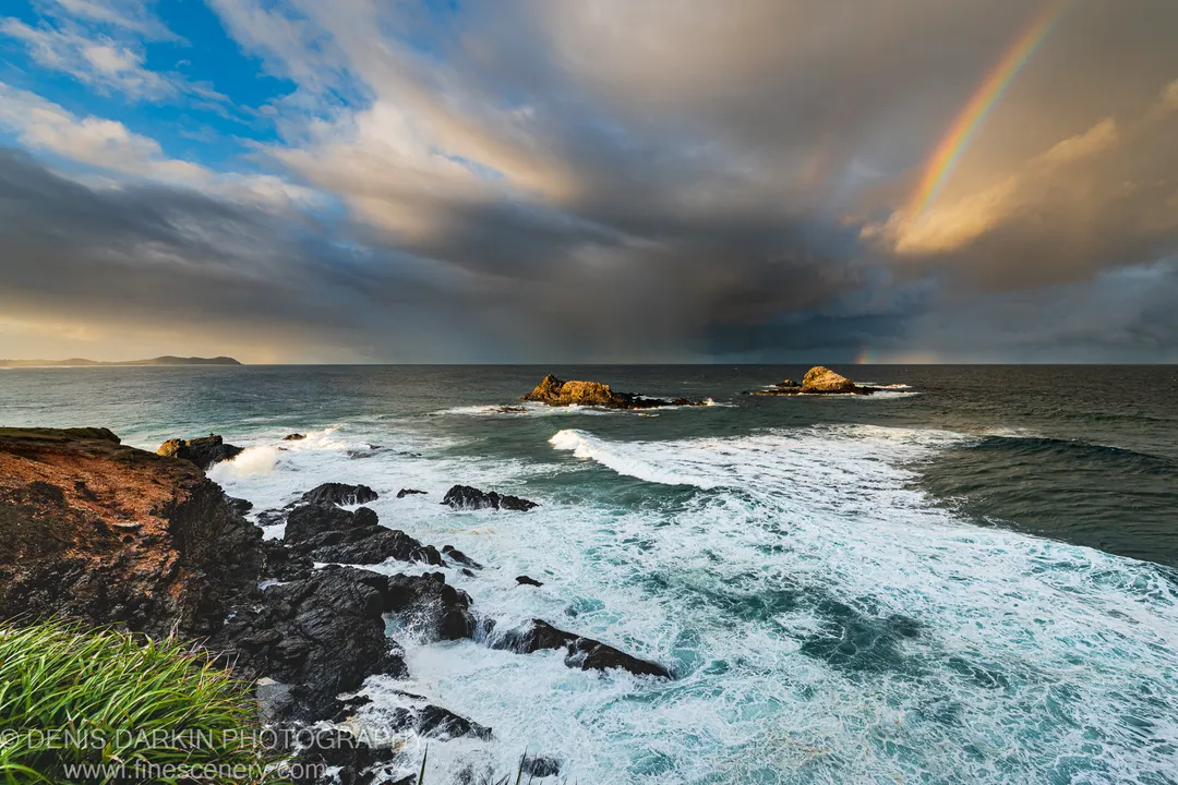 A large wave breaks against dark volcanic rocks as a rainbow arcs through storm clouds above the New South Wales coast.