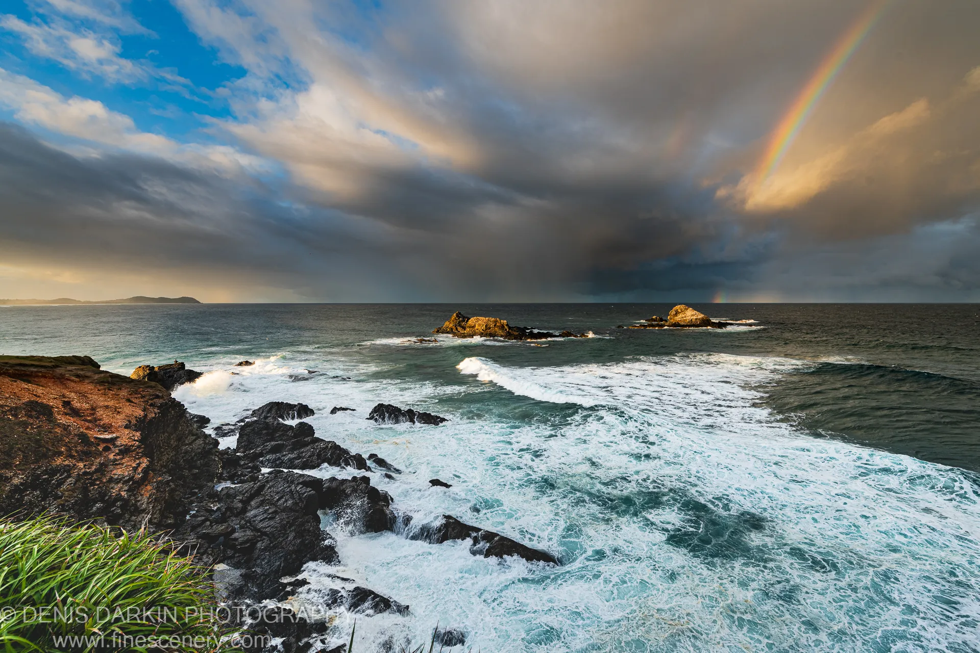 A large wave breaks against dark volcanic rocks as a rainbow arcs through storm clouds above the New South Wales coast.