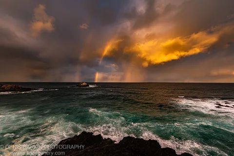 Dark rocks at Byron Bay below a gold-backlit storm sky with faint rainbow arcs near the horizon and waves in the middle ground.