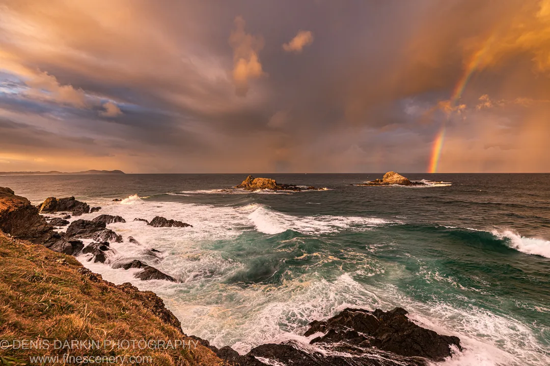 Warm golden-hour light falls on a rugged coastal shelf as a rainbow appears through heavy storm clouds on the horizon.