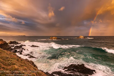 Warm golden-hour light falls on a rugged coastal shelf as a rainbow appears through heavy storm clouds on the horizon.