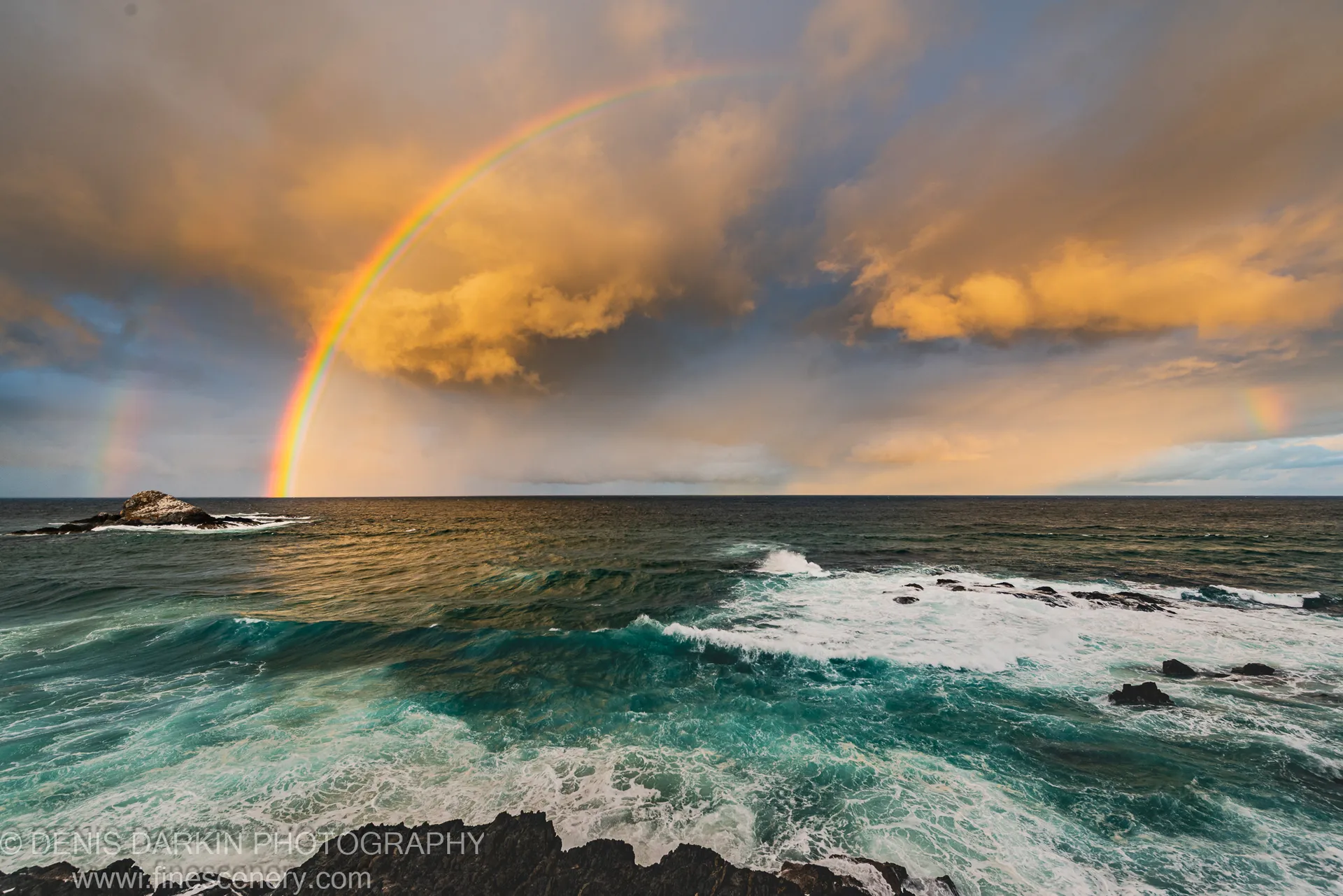A rainbow curves toward a solitary sea stack above rocky formations and crashing surf at Byron Bay, New South Wales.