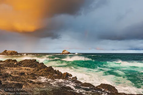Jagged coastal rocks at Byron Bay with swirling turquoise water, storm clouds overhead, and a band of golden light breaking through from the left.