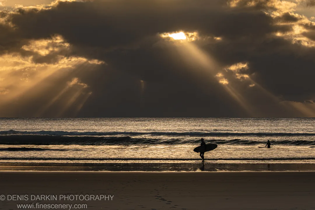 Crepuscular light shafts over the ocean at Byron Bay with two surfer silhouettes on the wet foreshore sand, winter morning.