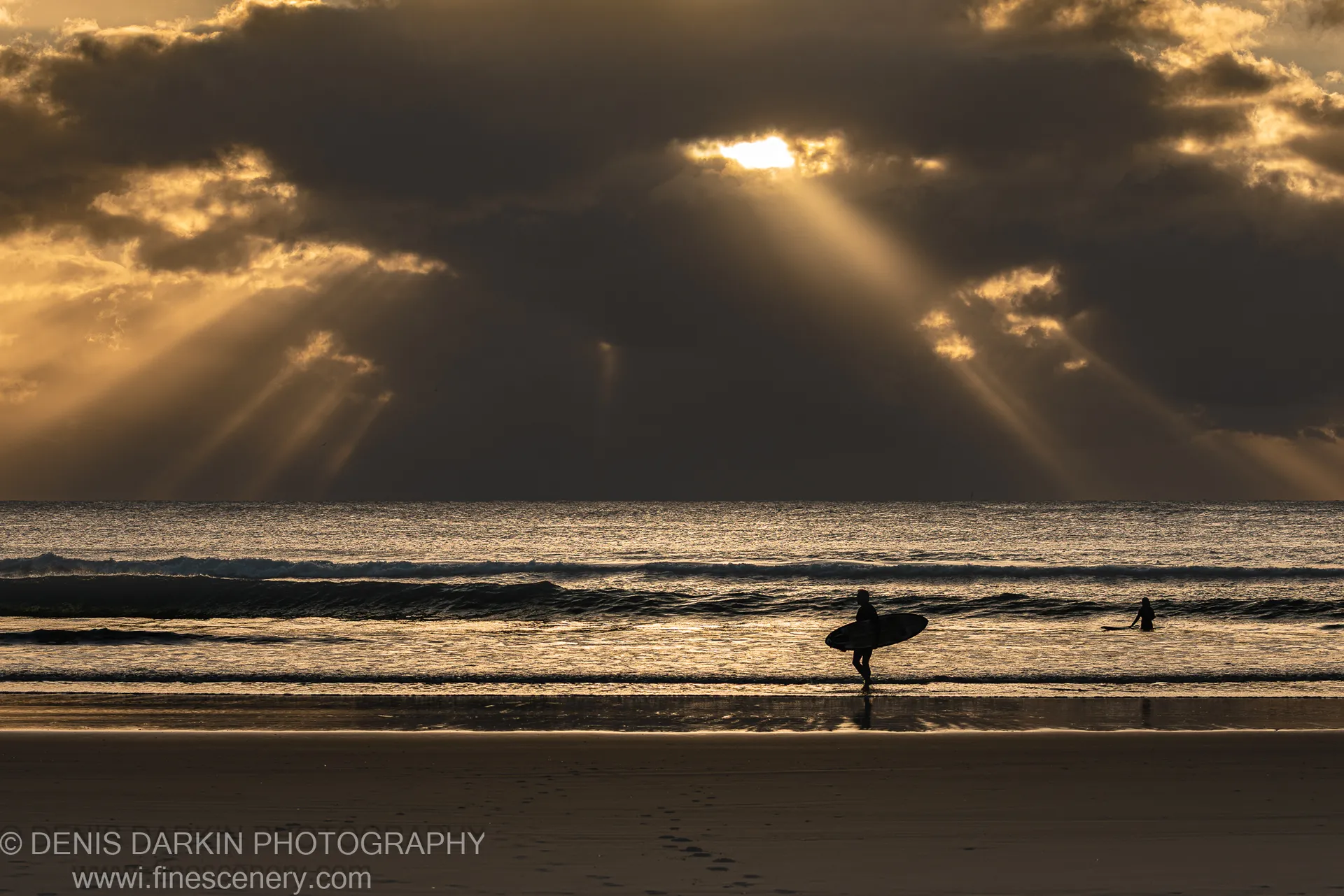 Crepuscular light shafts over the ocean at Byron Bay with two surfer silhouettes on the wet foreshore sand, winter morning.