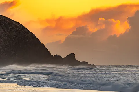 A dark rocky headland in silhouette against an orange-lit dawn sky at Byron Bay, waves rolling below through sea spray. Winter.