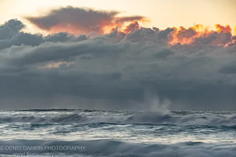 A massive bank of dark storm clouds over Byron Bay, lit from below at sunrise, with choppy surf and spray in the foreground.