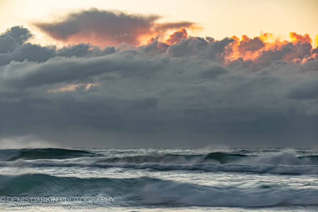 Long-exposure waves at Byron Bay, with a heavily backlit cloud layer showing orange and pink highlights above the dark ocean surface. Winter dawn.