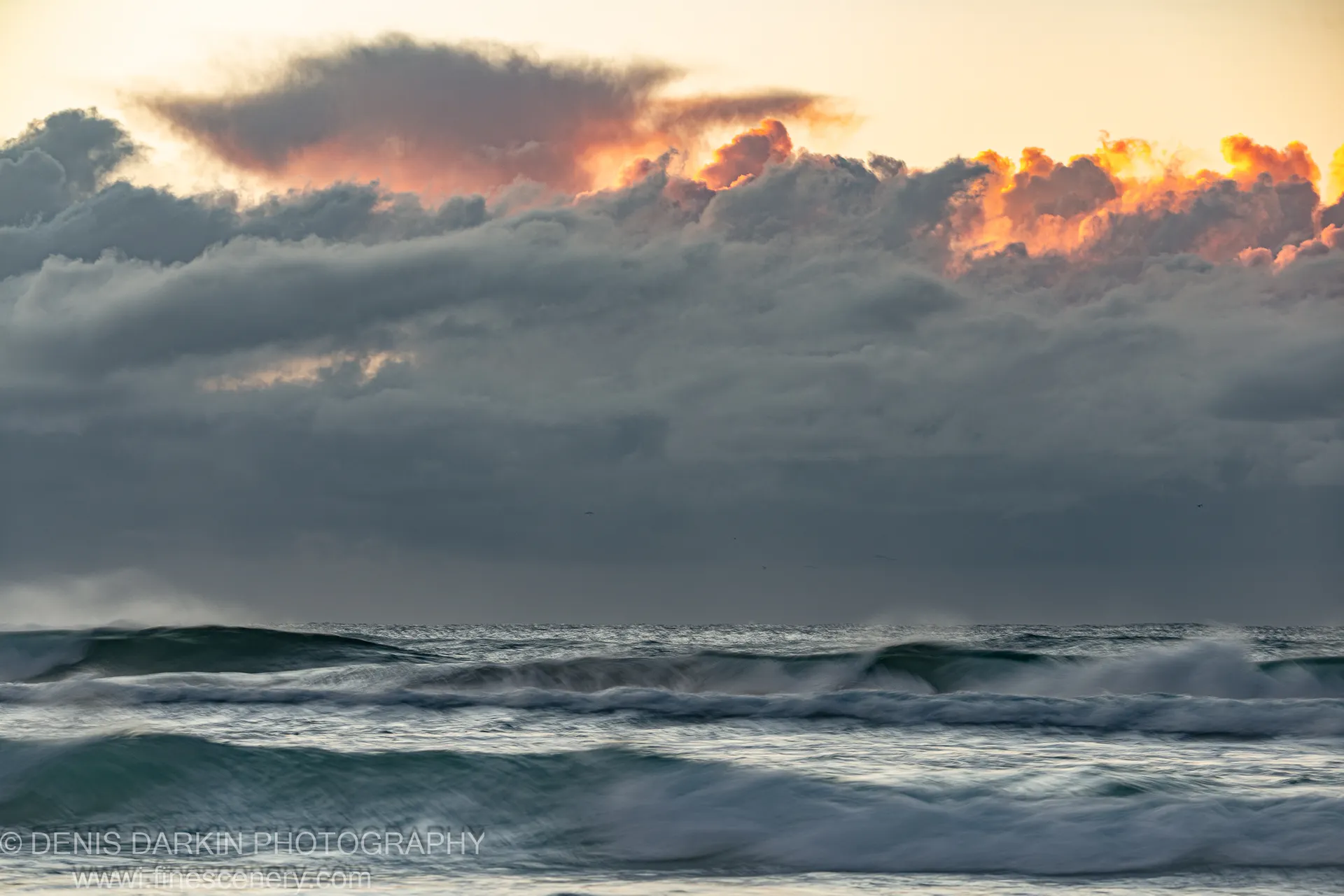Long-exposure waves at Byron Bay, with a heavily backlit cloud layer showing orange and pink highlights above the dark ocean surface. Winter dawn.