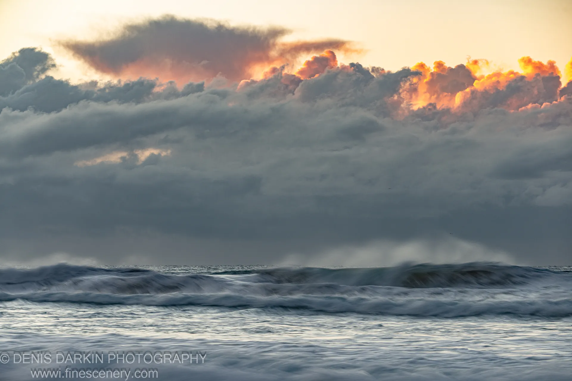 Motion-blurred waves at Byron Bay below a storm cloud bank illuminated in orange and pink tones, winter dawn over the New South Wales coast.