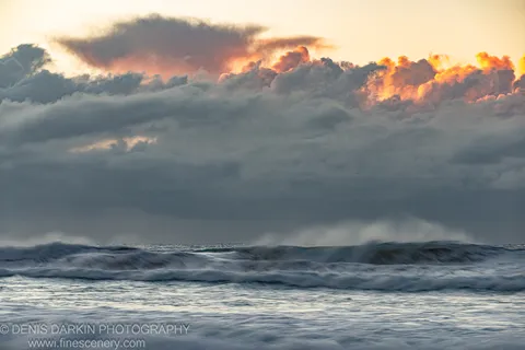 Motion-blurred waves at Byron Bay below a storm cloud bank illuminated in orange and pink tones, winter dawn over the New South Wales coast.