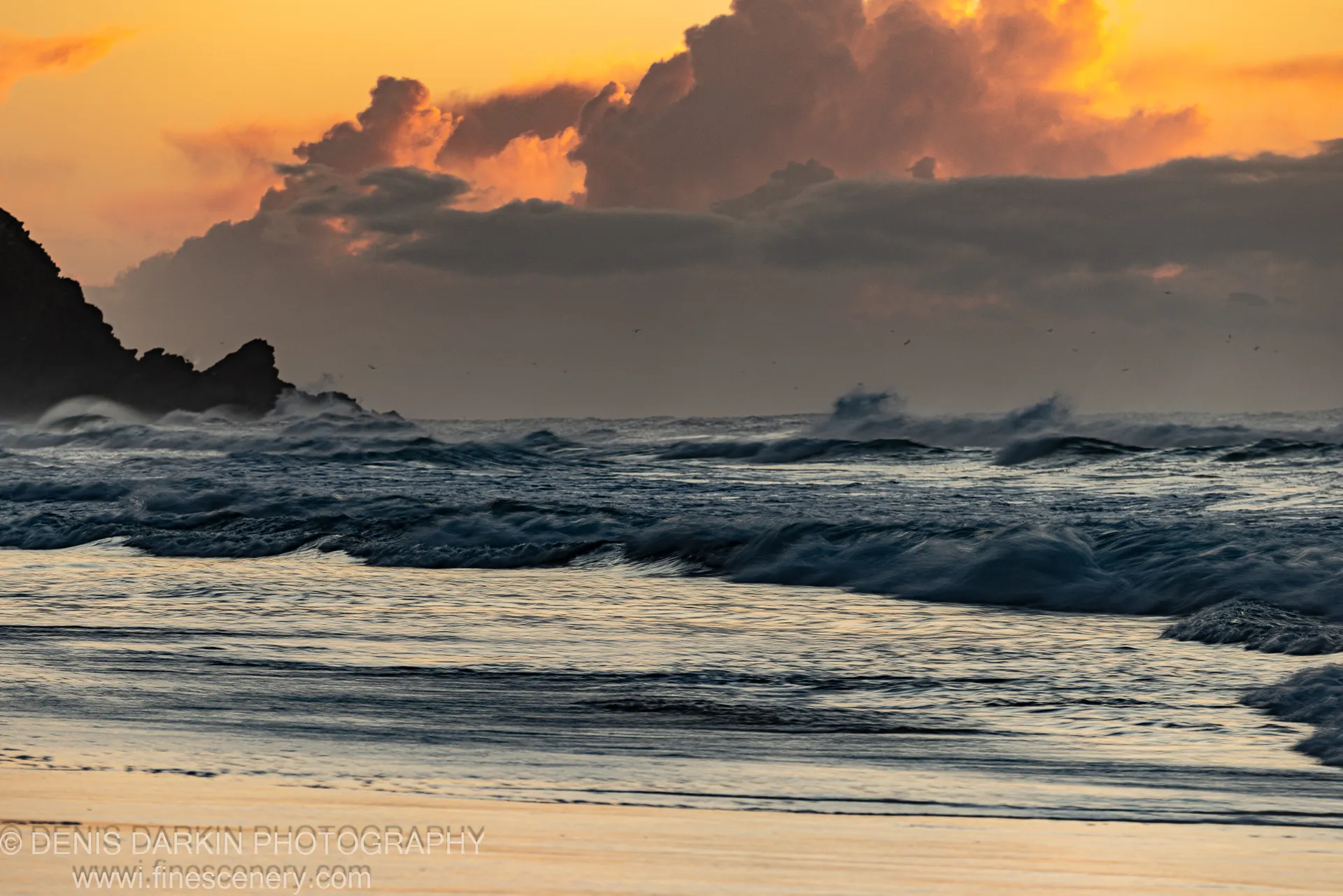 Deep blue swells at a sandy Byron Bay shore at dawn, silhouetted rock formations on the left under an orange-lit sky. Winter.