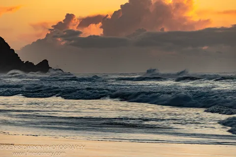 Deep blue swells at a sandy Byron Bay shore at dawn, silhouetted rock formations on the left under an orange-lit sky. Winter.