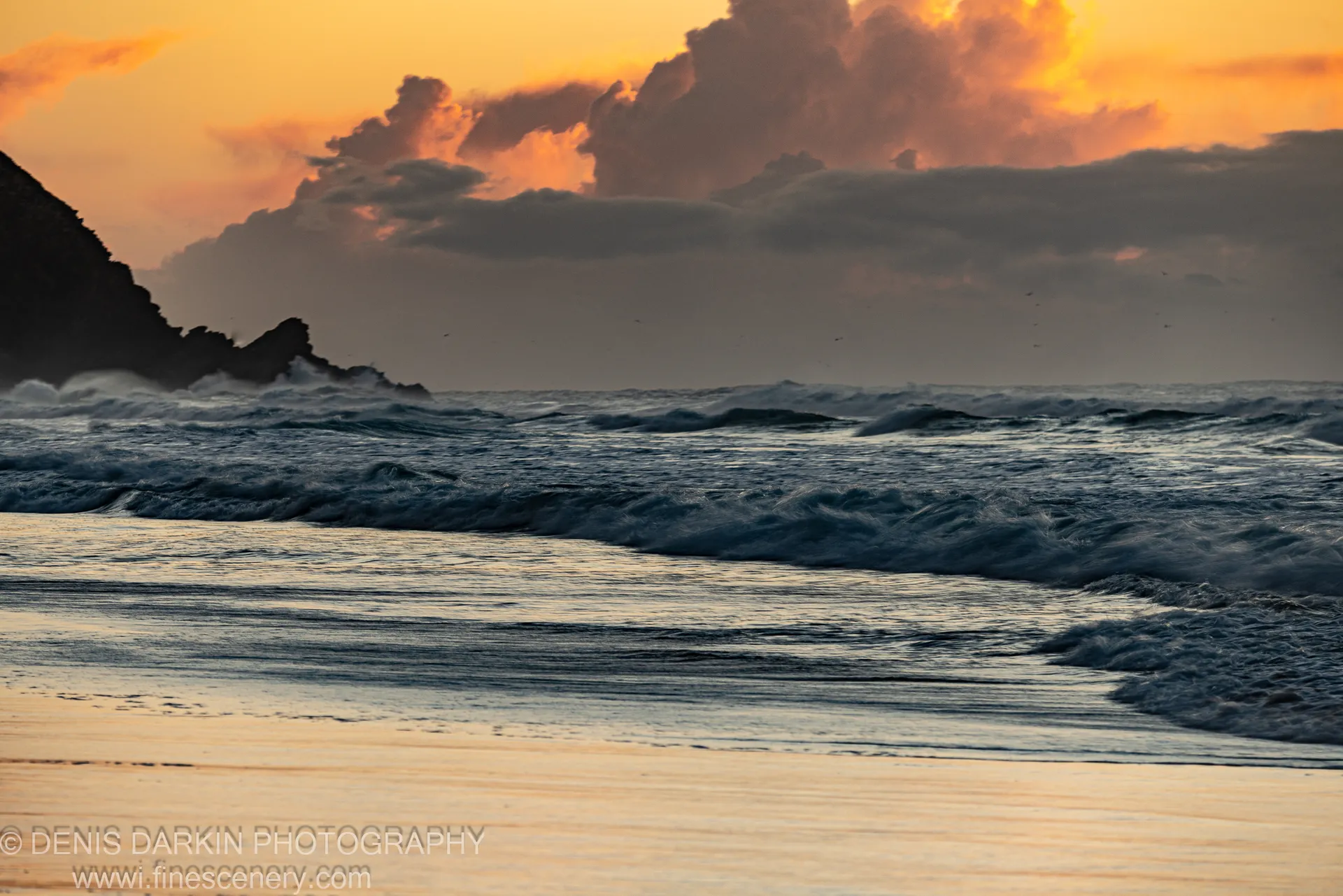 A dark silhouetted headland at Byron Bay with white spray crashing against it, contrasting with deep blue water and orange-lit clouds above. Winter dawn.