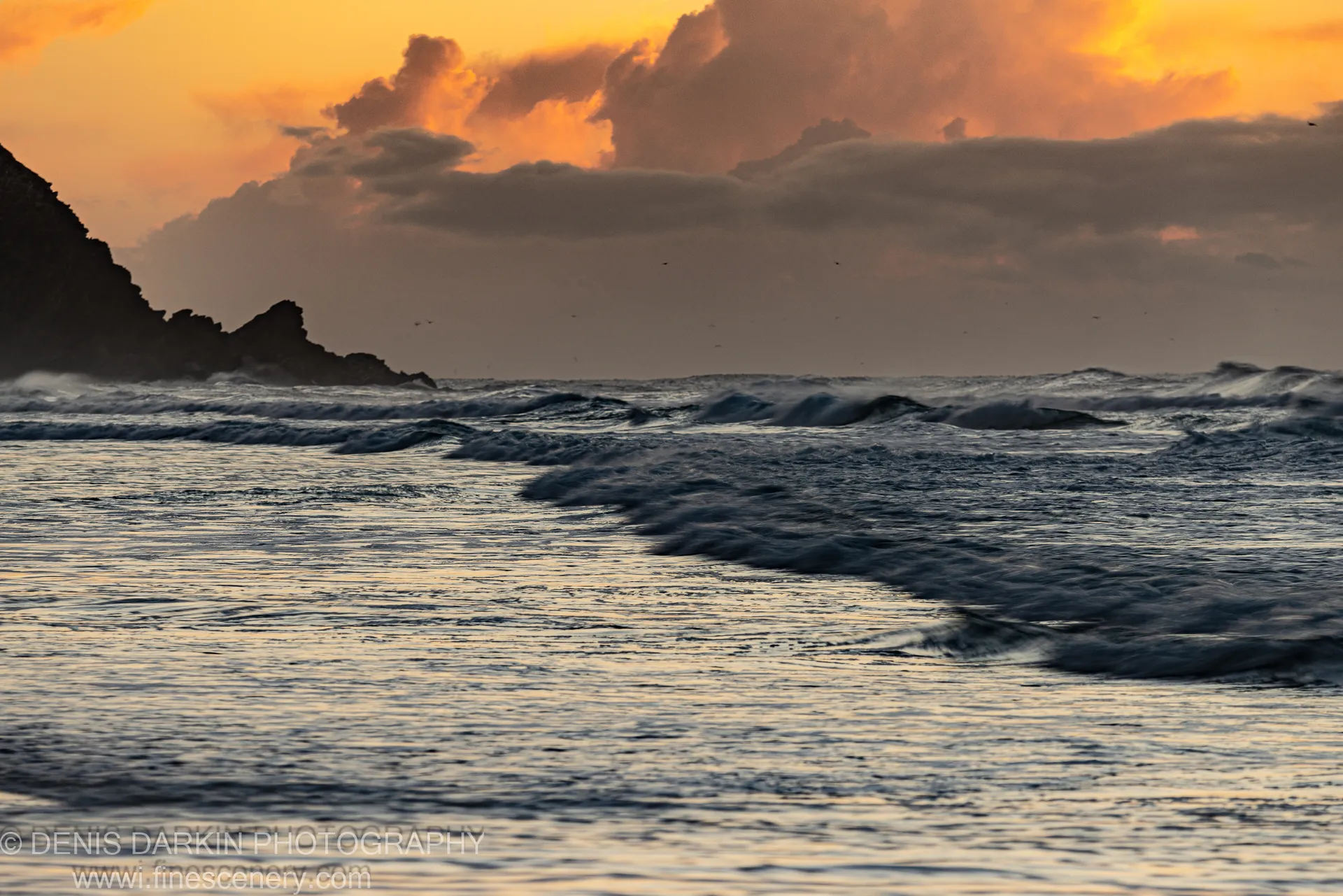 Silhouetted headland at Byron Bay with golden dawn reflections across the ocean, a foam path leading into the distance. Winter.