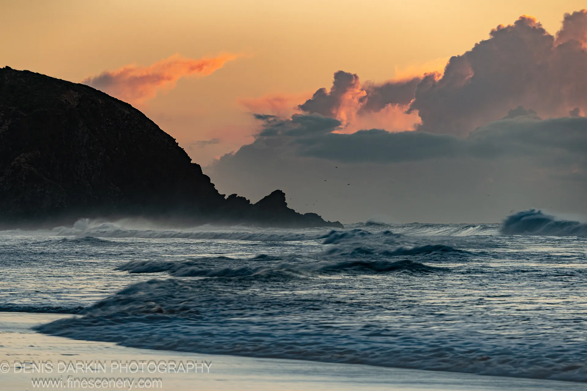 A silhouetted headland to the left, surf rolling onto wet sand at dawn, cloud undersides lit amber over Byron Bay, New South Wales.