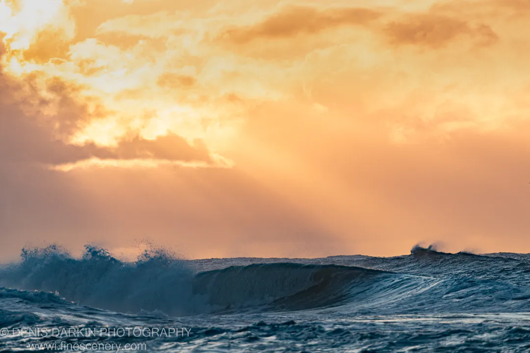 Diagonal sunbeams extend through clouds above a large incoming wave at Byron Bay, NSW, the sky filled with amber light at dawn.