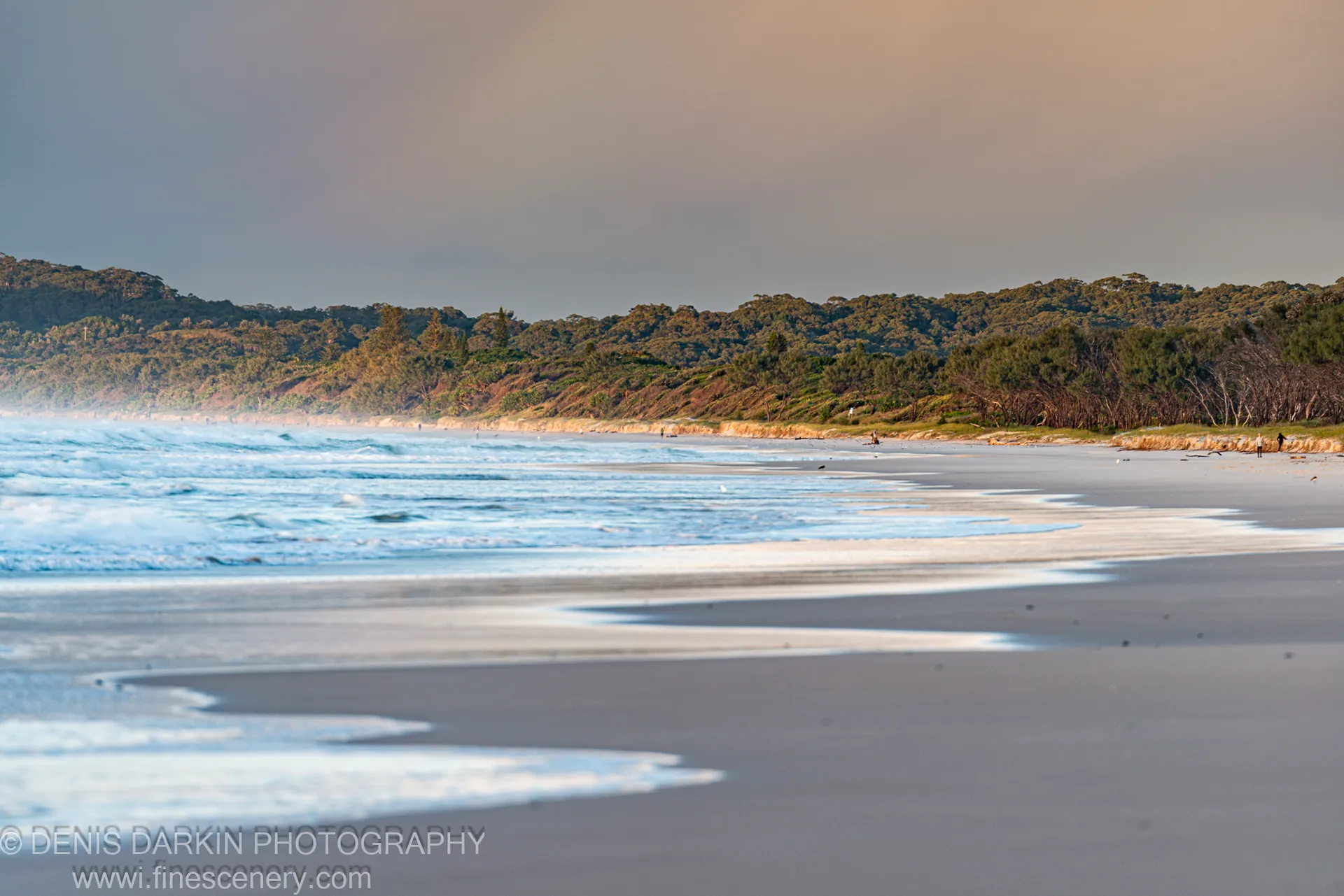 Rolling surf washes across wet sand toward a distant tree-covered headland at Byron Bay, NSW, the sky muted in early morning light.