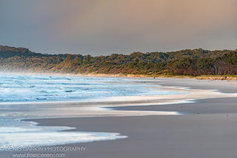 Rolling surf washes across wet sand toward a distant tree-covered headland at Byron Bay, NSW, the sky muted in early morning light.