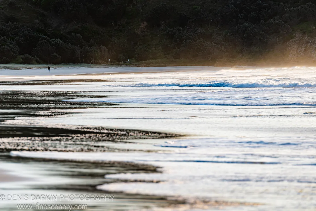 A solitary figure walks the tide line at Byron Bay, NSW, the beach stretching in horizontal bands of wet sand toward a wooded headland.