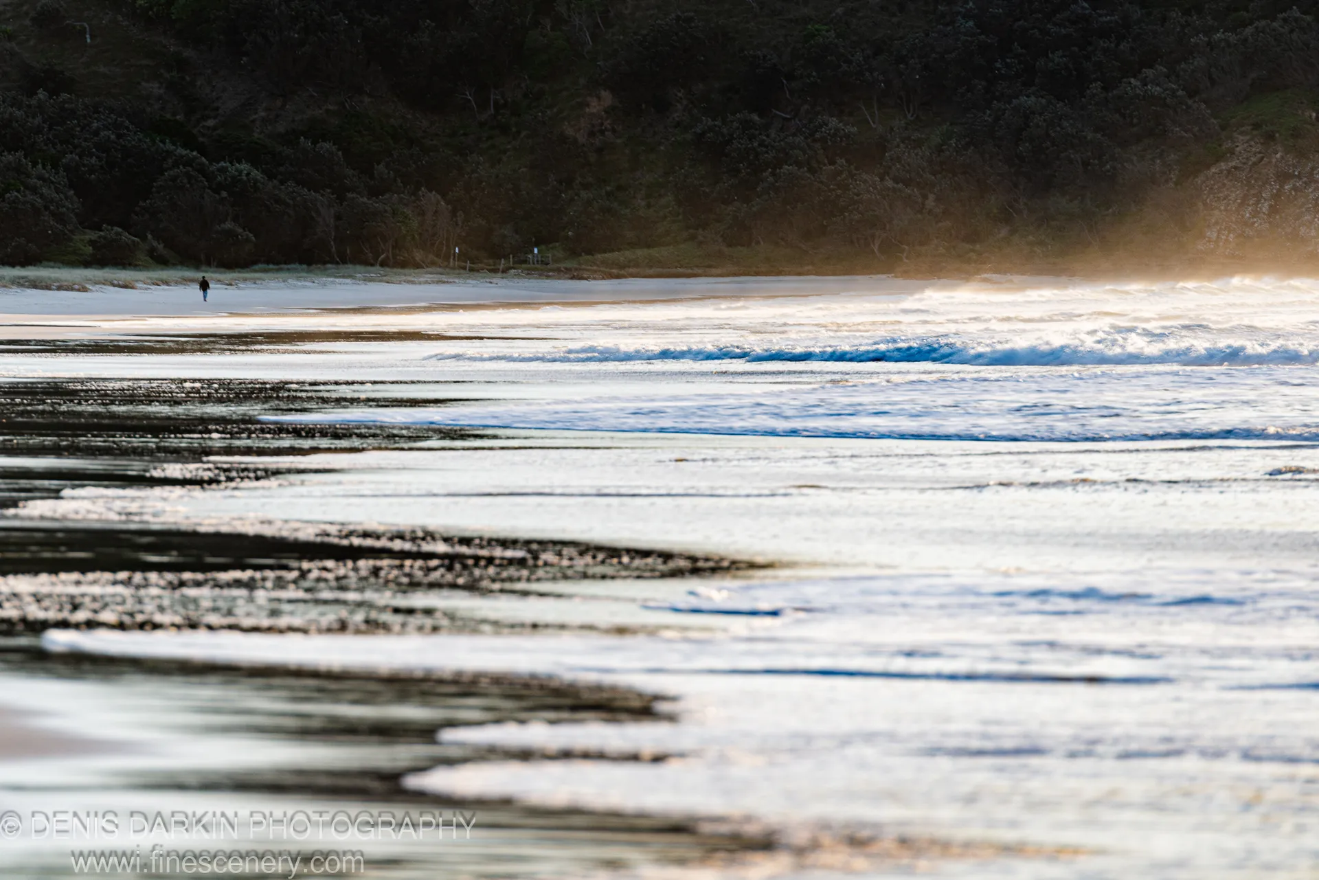 A solitary figure walks the tide line at Byron Bay, NSW, the beach stretching in horizontal bands of wet sand toward a wooded headland.
