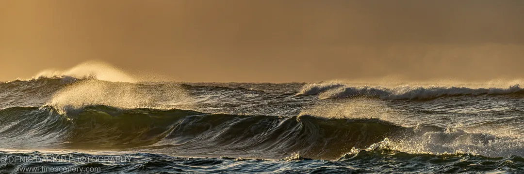 Telephoto compression stacks rolling ocean swells in morning light at Byron Bay, NSW, fine spray catching warm amber color at the breaking crests.
