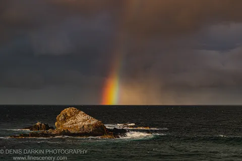 A weathered coastal rock formation in warm sunlight stands against a grey storm sky with a rain curtain and faint rainbow behind it.