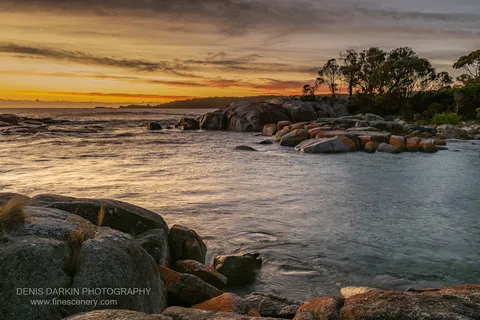 Long-exposure water flows around orange lichen boulders in a coastal inlet at Bay of Fires, Tasmania, under an amber and grey morning sky.