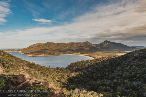 Distant mountain ridges catch warm afternoon light above the deep blue waters of Wineglass Bay, with scrub-covered slopes in the foreground.