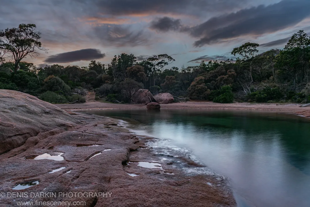 Rust-toned granite lines the Coles Bay shoreline on a cloudy late-autumn morning, with pink light visible in the cloud formations above.