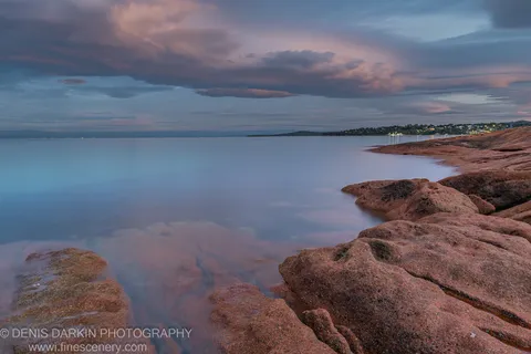 Pink granite boulders at Coles Bay emerge through long-exposure water on a late-autumn morning, a distant shore visible beneath streaked cloud.