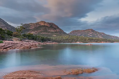 Reddish granite boulders at Coles Bay stand before smooth long-exposure water, a mountain peak visible above forested hills on an overcast morning.