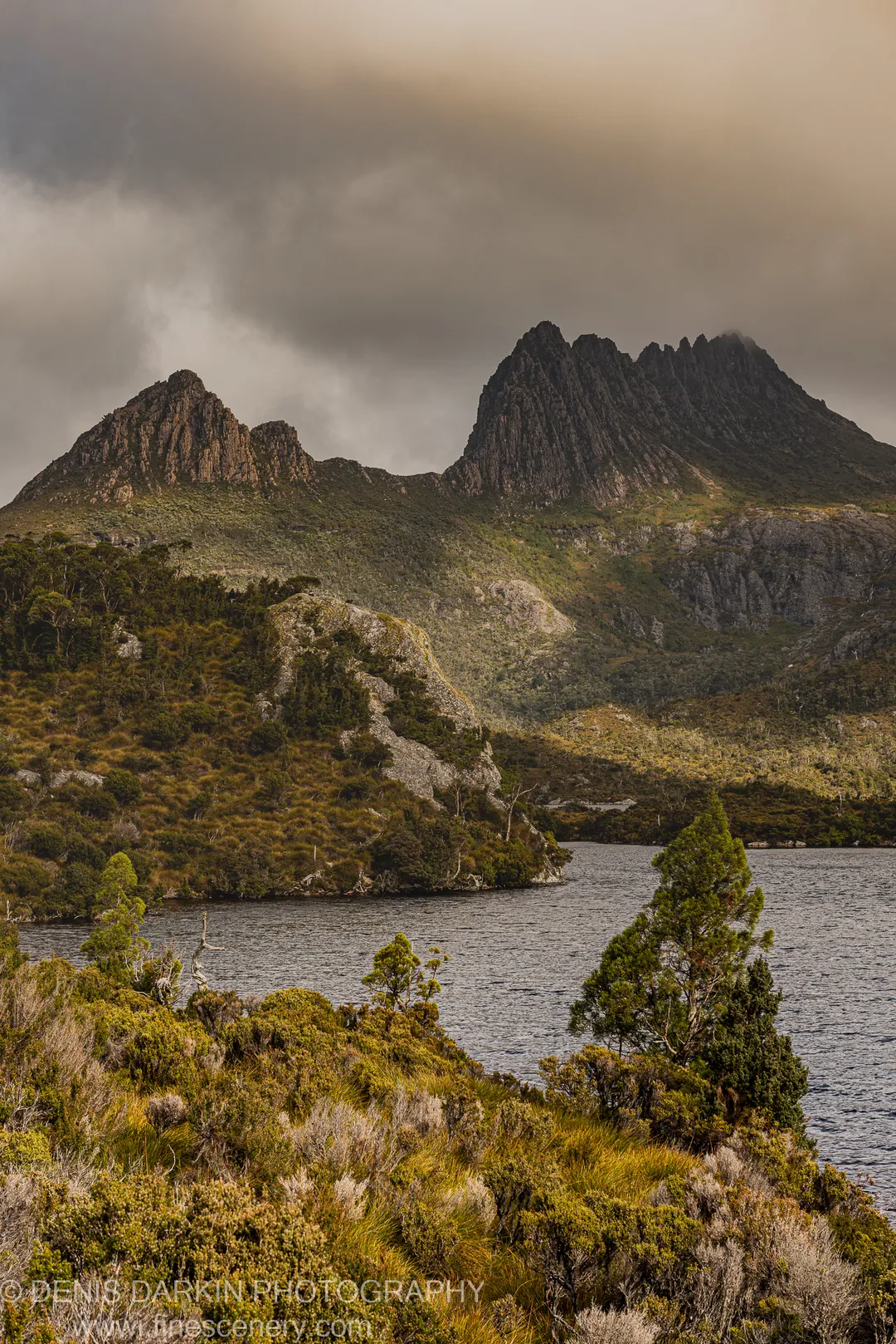 Heavy storm clouds loom over the dolerite twin peaks at Cradle Mountain, with ochre tussock scrub and a dark alpine lake in the foreground.