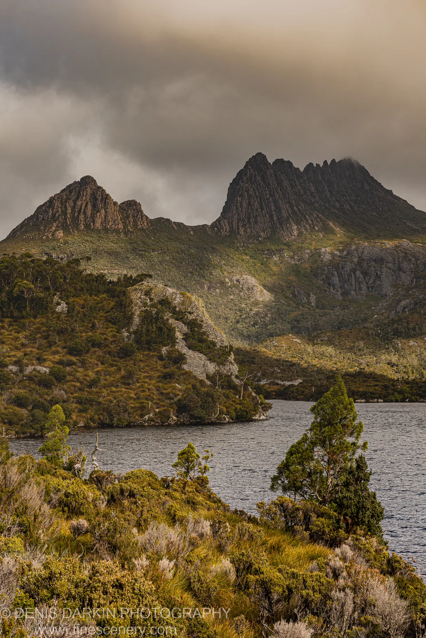 Heavy storm clouds loom over the dolerite twin peaks at Cradle Mountain, with ochre tussock scrub and a dark alpine lake in the foreground.
