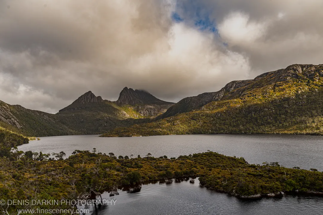 Soft light breaks through heavy clouds over dolerite peaks at Cradle Mountain, with dark still water reflecting the buttongrass heath shoreline below.