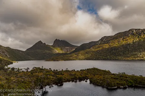 Soft light breaks through heavy clouds over dolerite peaks at Cradle Mountain, with dark still water reflecting the buttongrass heath shoreline below.