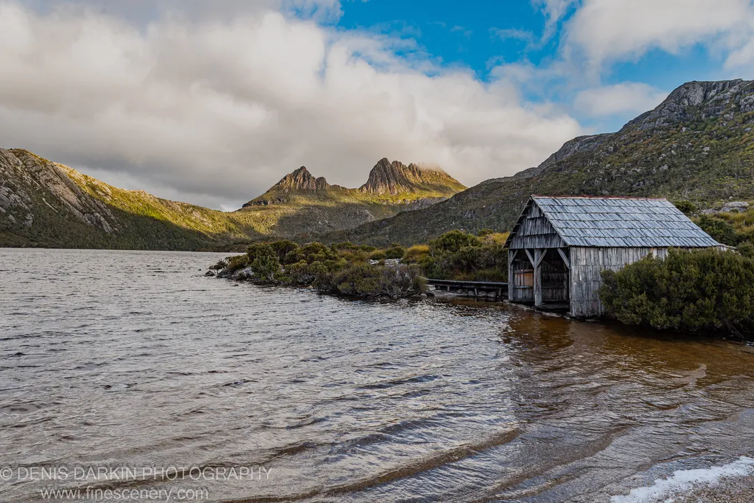 A weathered wooden boathouse on the Cradle Mountain lakeshore, with jagged ridges catching warm afternoon light and rippling water below.