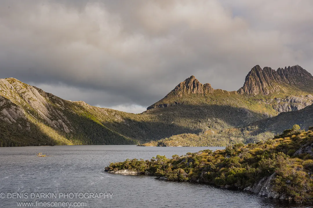 Rocky peaks dusted with scrub catch directional light above a calm alpine lake at Cradle Mountain, Tasmania.