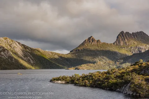 Rocky peaks dusted with scrub catch directional light above a calm alpine lake at Cradle Mountain, Tasmania.