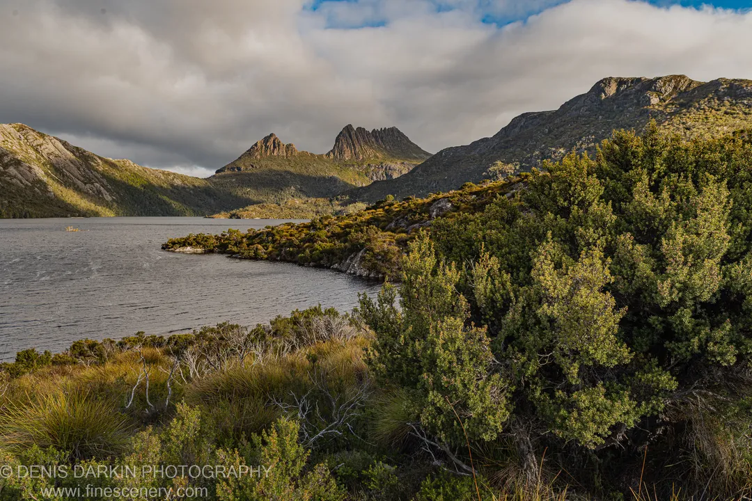 A break in heavy cloud sends warm light across the jagged ridges at Cradle Mountain, shadowed lower slopes meeting dark rippled water below.