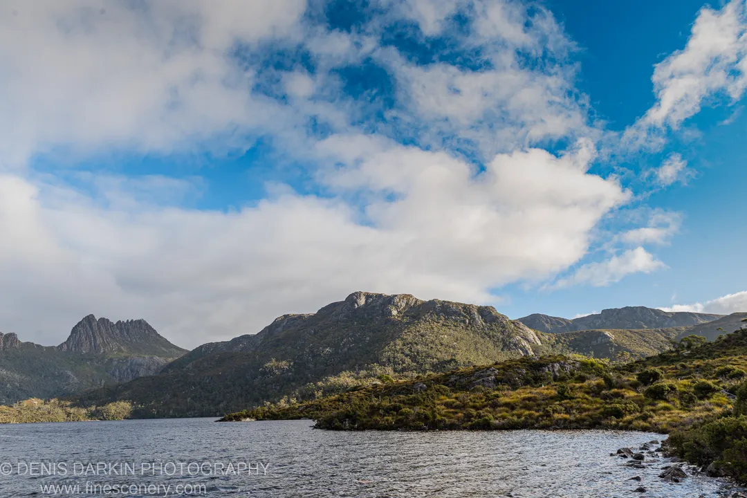 Textured ripples on a dark lake surface at Cradle Mountain, sunlit granite peaks and scattered clouds above scrub-covered slopes in the distance.
