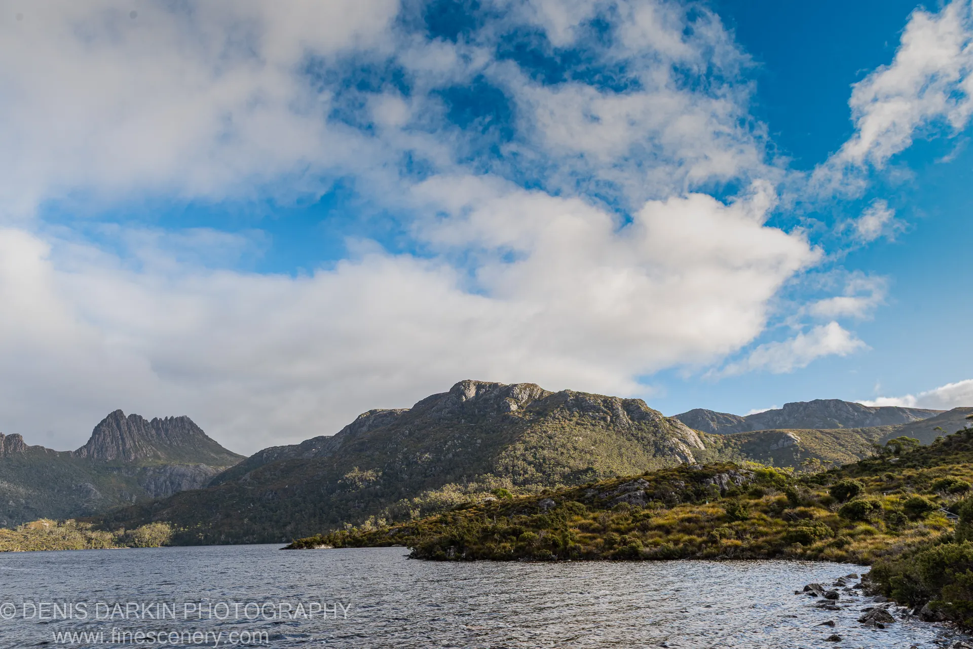 Textured ripples on a dark lake surface at Cradle Mountain, sunlit granite peaks and scattered clouds above scrub-covered slopes in the distance.