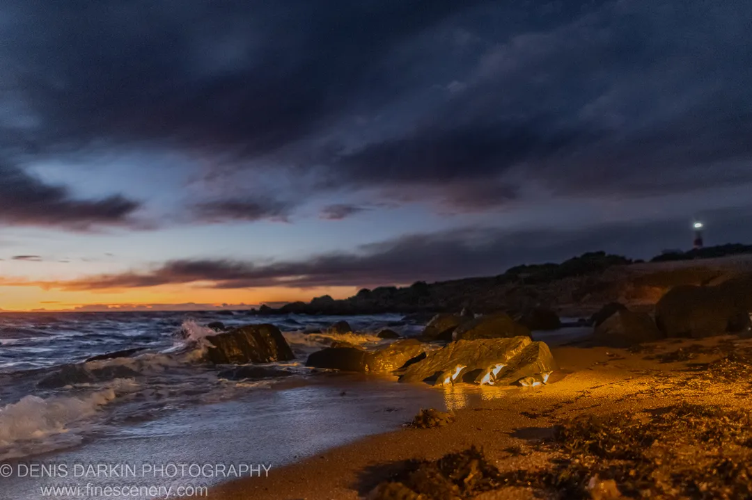 Warm amber light on wet rocks and sand at Lillico Beach, heavy clouds over the darkening ocean, a lighthouse on the far shore, Tasmania.