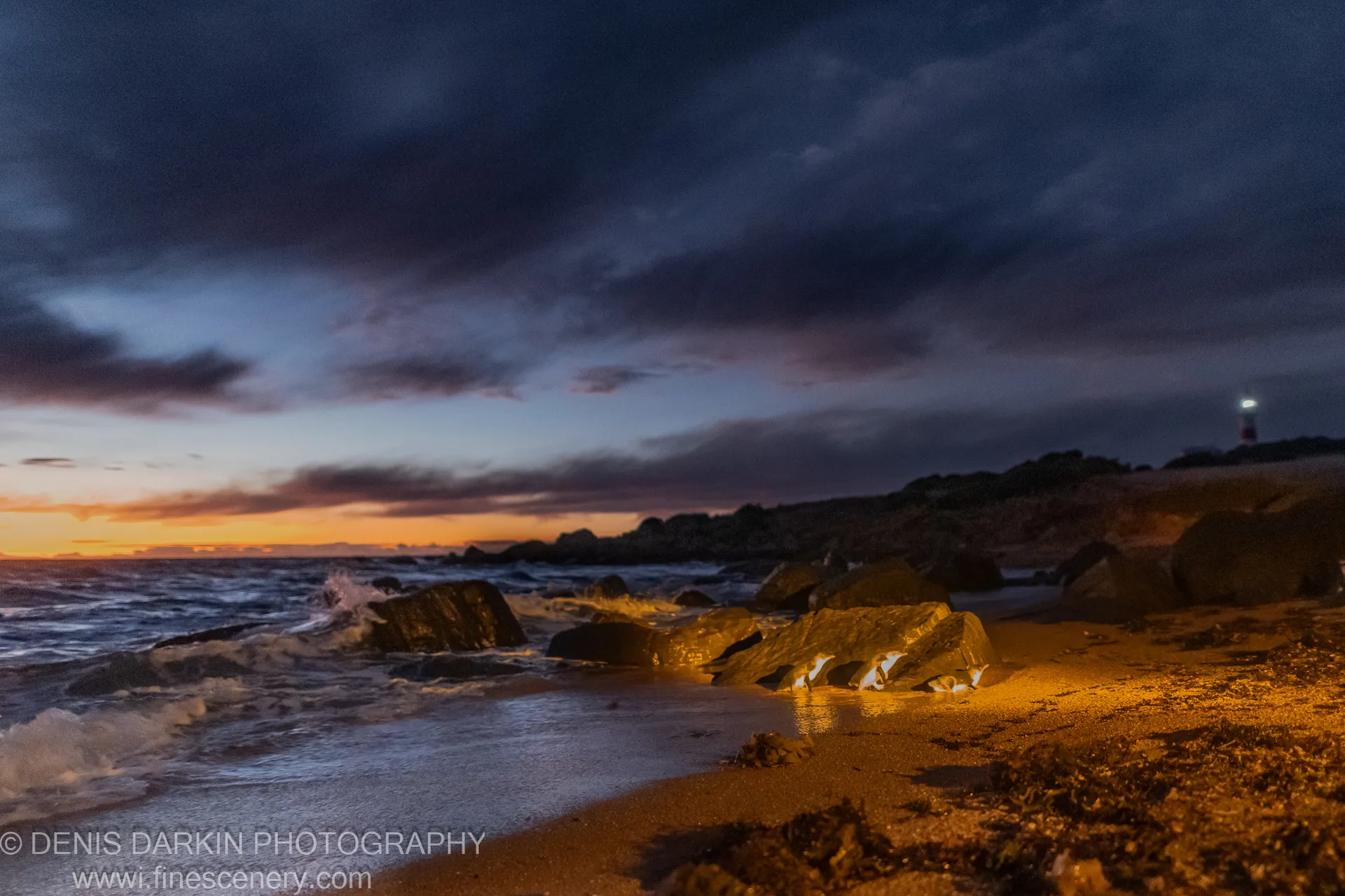 Warm amber light on wet rocks and sand at Lillico Beach, heavy clouds over the darkening ocean, a lighthouse on the far shore, Tasmania.