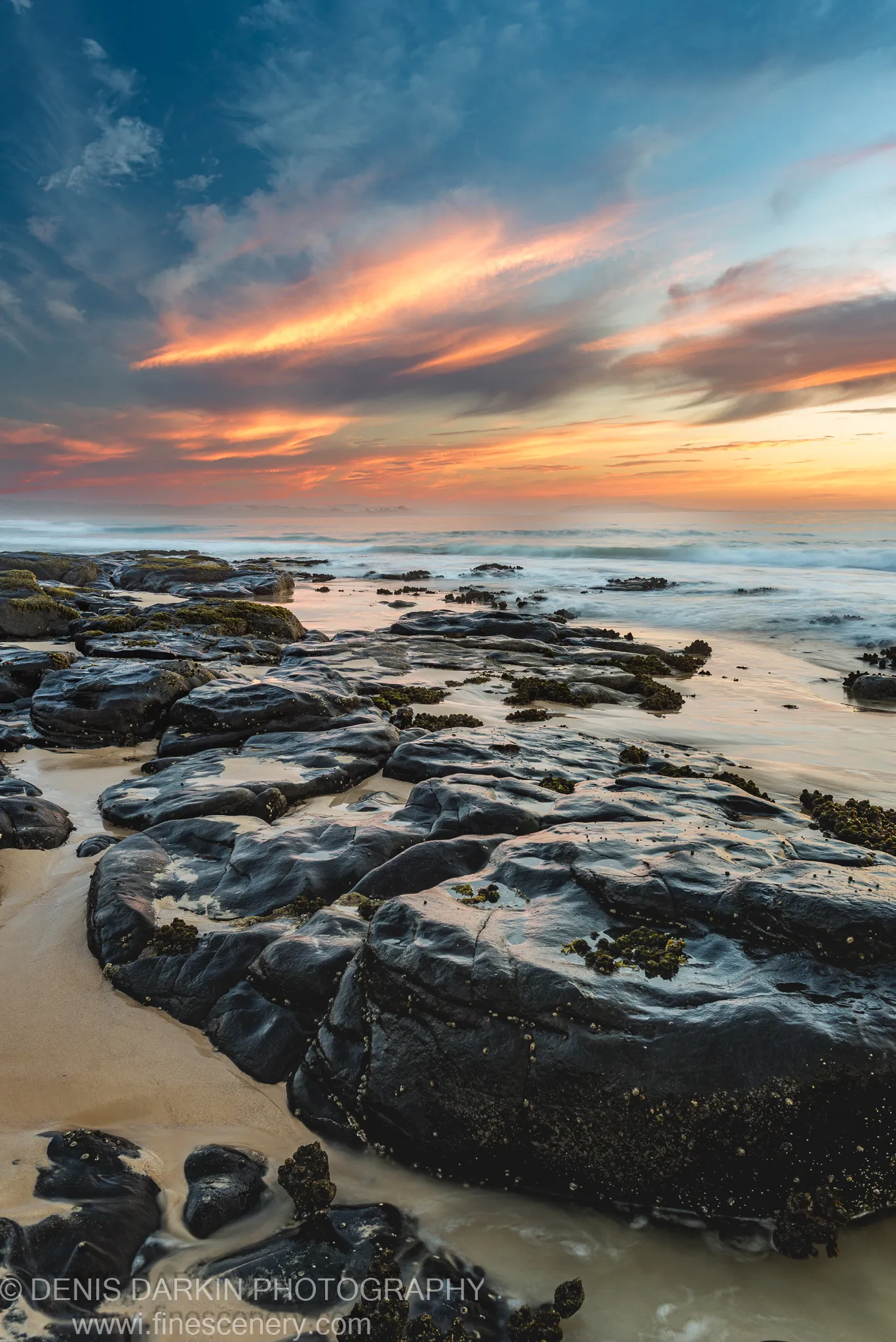 Marine-growth-covered rock platform on the New South Wales coast, with long-exposure surf and orange sunrise clouds above the horizon.