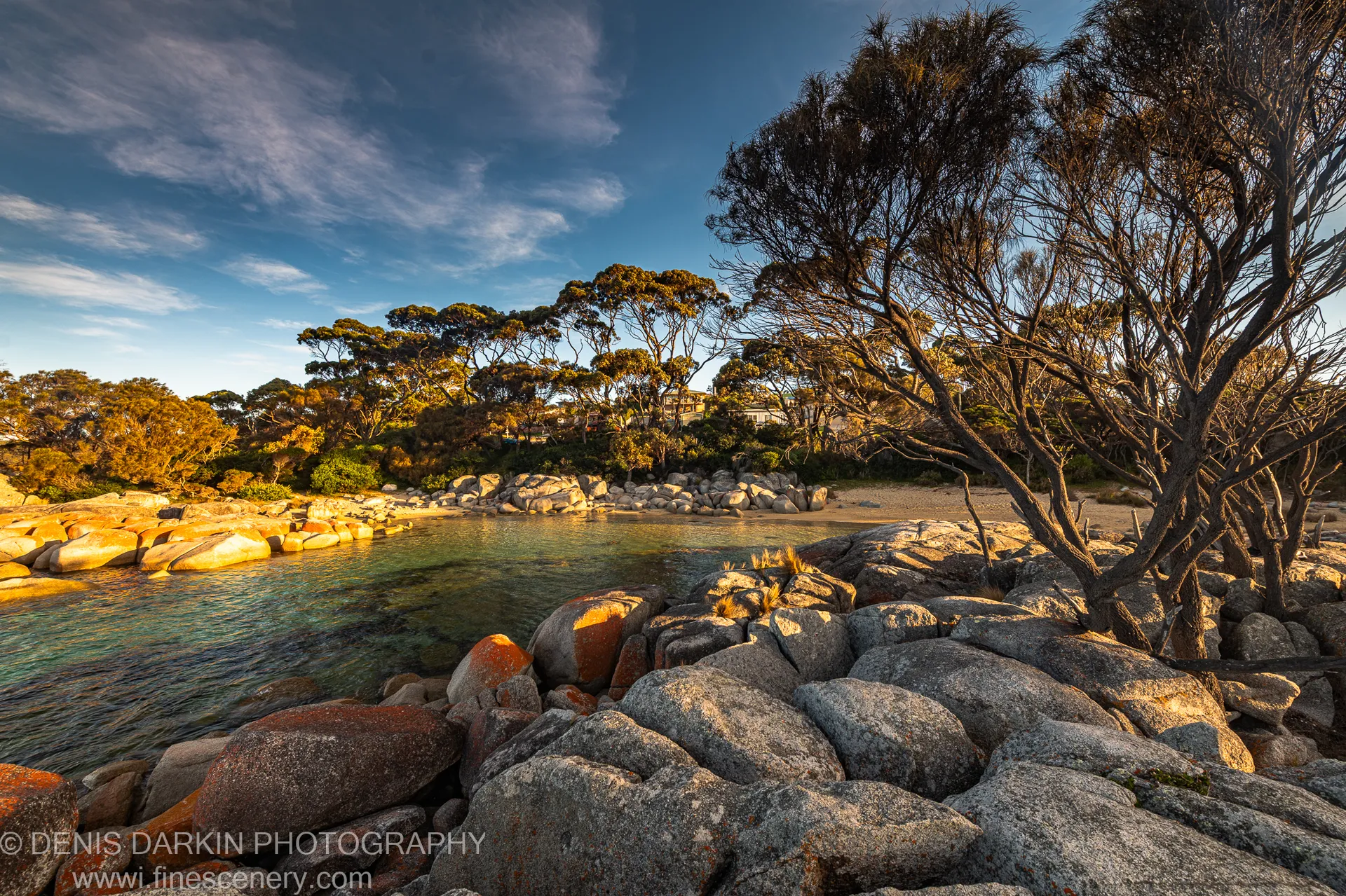 Rounded granite boulders in warm light at a tidal inlet, Bay of Fires, eucalyptus branches above the water, a distant shore under blue sky, Tasmania.