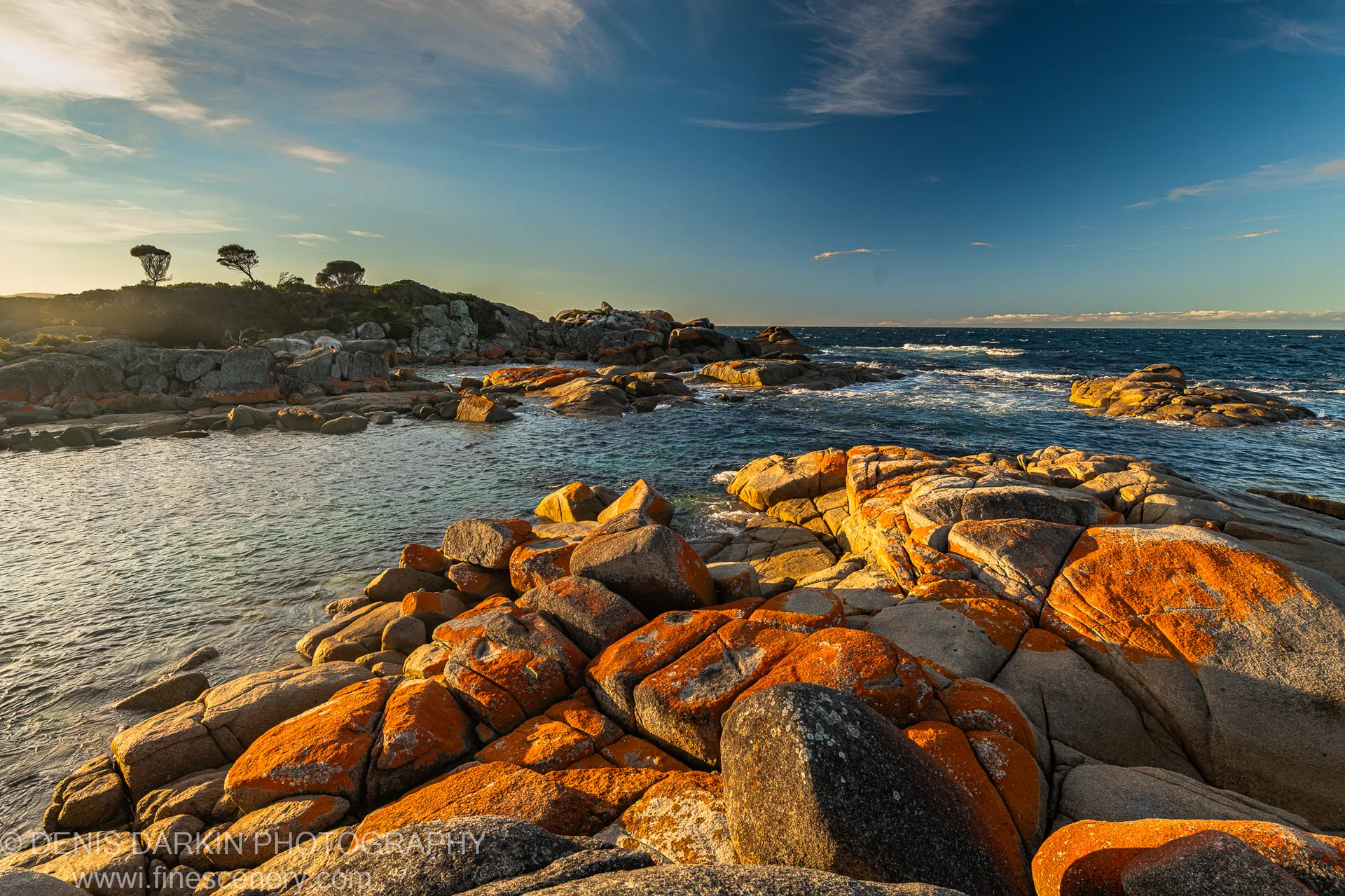 Rust-orange lichen-covered granite rocks in low evening light, with wispy clouds above the distant headland at Bay of Fires, Tasmania.